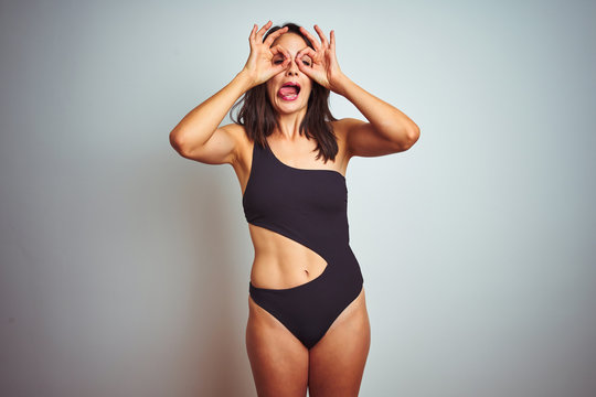 Beautiful woman wearing bikini swimwear over white isolated background doing ok gesture like binoculars sticking tongue out, eyes looking through fingers. Crazy expression.