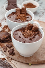 Two mugs of a hot chocolate with chocolate sticks on a wooden board