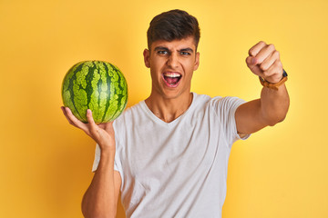 Young indian shopkeeper man holding watermelon standing over isolated yellow background annoyed and frustrated shouting with anger, crazy and yelling with raised hand, anger concept