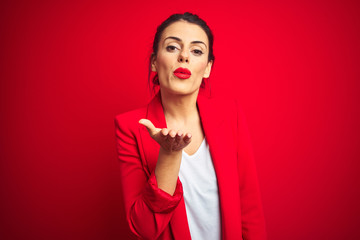 Young beautiful business woman standing over red isolated background looking at the camera blowing a kiss with hand on air being lovely and sexy. Love expression.
