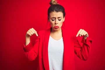 Young beautiful business woman standing over red isolated background Pointing down looking sad and upset, indicating direction with fingers, unhappy and depressed.