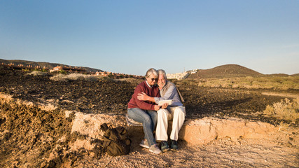 Fototapeta premium Attractive couple of senior people sitting together on the cliff in front to the sea. Looking at tablet and smiling. Sunrise and arid landscape as background