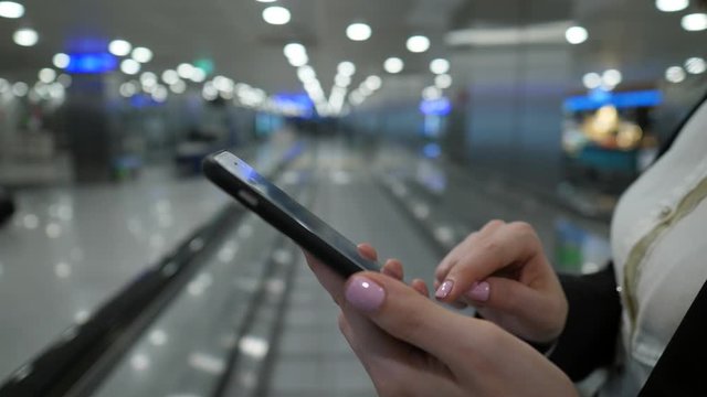 Woman texting using smartphone, closeup shot of mobile device in hands. Lady stay on moving travolator, blurred airport hall on seen background.