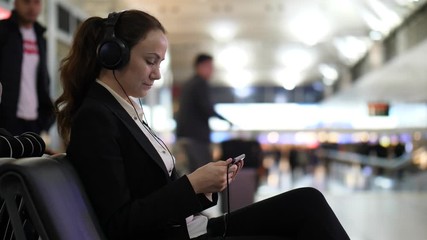 Business woman rest at airport at evening time, watch film on smartphone, listen sounds by headphones. Blurred background, silhouettes of passengers, people walk to boarding gates at airport