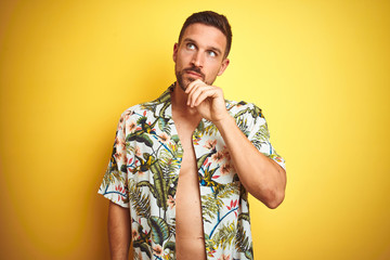 Handsome man wearing summer hawaiian flowers shirt over yellow isolated background with hand on chin thinking about question, pensive expression. Smiling with thoughtful face. Doubt concept.