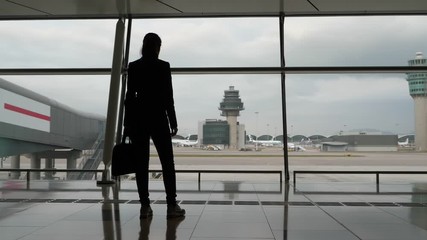 Thoughtful woman stay against terminal window, full length silhouetted shot. Black figure of lady with laptop bag in hands standing still and looking out to airport apron and taxiing field