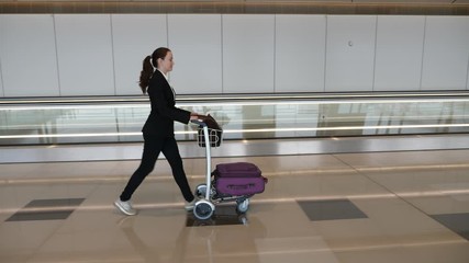 Passenger woman walk at airport passage, push ahead small cart loaded with hand luggage, tracking camera motion. Lady wear informal attire, go alone beside empty travolator.