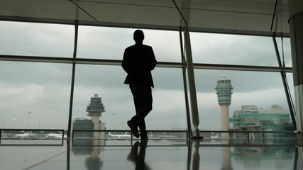 Traveler man walk to full height glass wall at terminal and look out to airport taxiways, stay with hands on hips. Black silhouette of man in casual suit against large window.