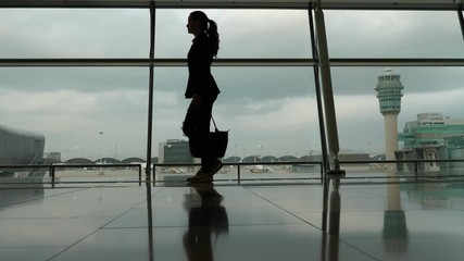 Woman silhouette against full height window, she walk at arrival passage of international terminal. Black figure of passenger go by, control tower and airport taxiing field seen outdoors