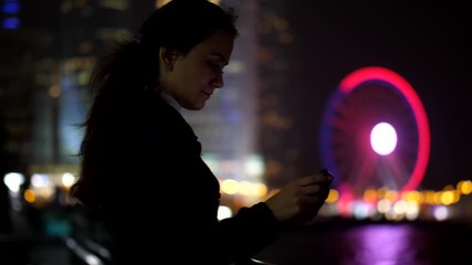 Woman browse photographs on smartphone, stay at Hong Kong Island embankment, night time. Illuminated ferris wheel seen blurred on background, lady stare to mobile device screen