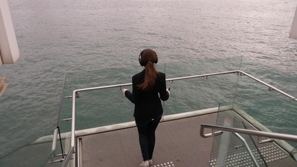 Happy woman enjoy music and beautiful waterfront, run downstairs and stay lean to glass railings, admire Victoria Harbour panorama. High angle slow motion shot, water surface seen on background