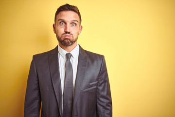 Young handsome business man over yellow isolated background puffing cheeks with funny face. Mouth inflated with air, crazy expression.