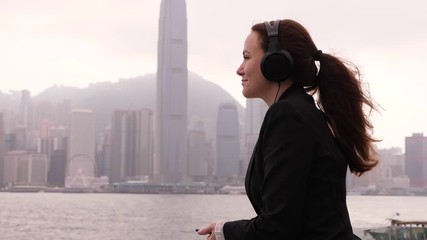 Woman listen music on headphones, stay at Victoria Harbour embankment, enjoy free time, admire beautiful Hong Kong city waterfront. Long tail hair fly in light breeze, half length shot of lady