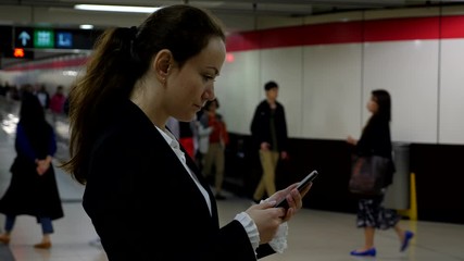 Lady watch smartphone, scroll web page, stay in metro passage, blurred background, some passengers pass by. Half length shot of woman standing with mobile device in hands