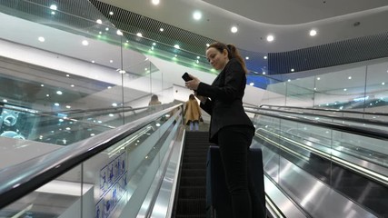 Woman chat using smartphone, stay at long escalator in shopping complex, travelling to upper floor with large trolley case. Low angle shot, bright modern atrium of Hong Kong emporium