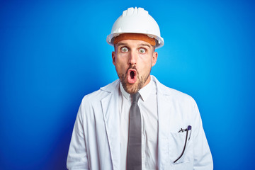 Young handsome engineer man wearing safety helmet over blue isolated background afraid and shocked with surprise expression, fear and excited face.