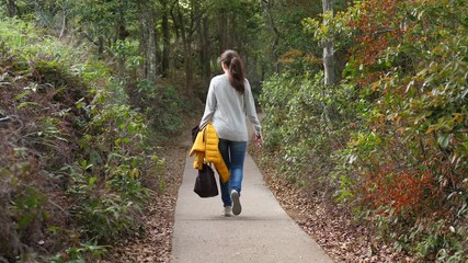 Woman walk away at empty path in country park, carry handbag and yellow jacket in hands, telephoto view from back. Empty trail at Lantau Island, surround with green verdure and bushes