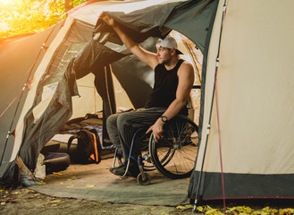 Disabled man resting in a campsite with friends. Wheelchair in the forest on the background of tents