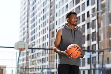 Waist up portrait of masculine African man holding basketball ball in court outdoors, copy space