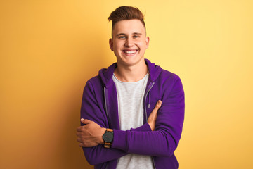 Young handsome man wearing purple sweatshirt standing over isolated yellow background happy face smiling with crossed arms looking at the camera. Positive person.