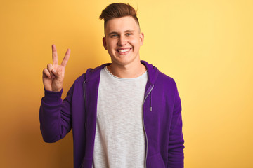 Young handsome man wearing purple sweatshirt standing over isolated yellow background smiling with happy face winking at the camera doing victory sign. Number two.