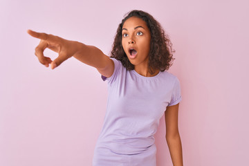 Young brazilian woman wearing t-shirt standing over isolated pink background Pointing with finger surprised ahead, open mouth amazed expression, something on the front