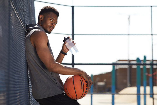 Side view portrait of handsome African-American man holding basketball ball looking at camera while drinking water in sports court outdoors, copy space