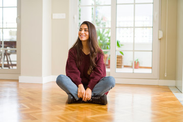 Young beautiful woman sitting on the floor at home looking away to side with smile on face, natural...