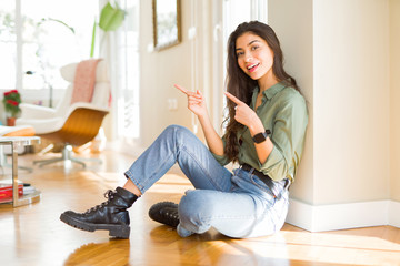 Young beautiful woman sitting on the floor at home smiling and looking at the camera pointing with two hands and fingers to the side.
