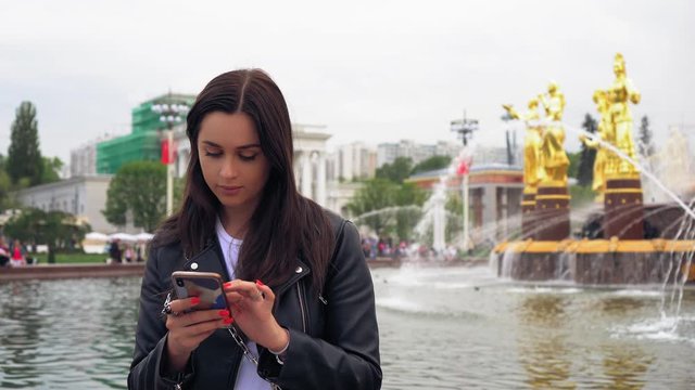 Beautiful girl chatting on a smartphone. Stylish clothes, red manicure. In the background is a beautiful fountain. Daytime, sunny weather.