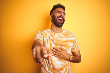 Young indian man wearing t-shirt standing over isolated yellow background laughing at you, pointing finger to the camera with hand over body, shame expression