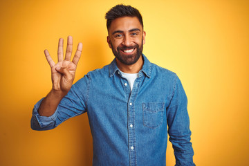 Young indian man wearing denim shirt standing over isolated yellow background showing and pointing up with fingers number four while smiling confident and happy.