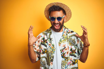 Indian man on vacation wearing floral shirt hat sunglasses over isolated yellow background...