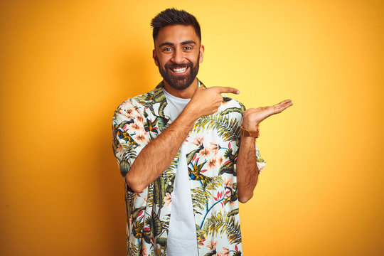 Young indian man on vacation wearing summer floral shirt over isolated yellow background amazed and smiling to the camera while presenting with hand and pointing with finger.