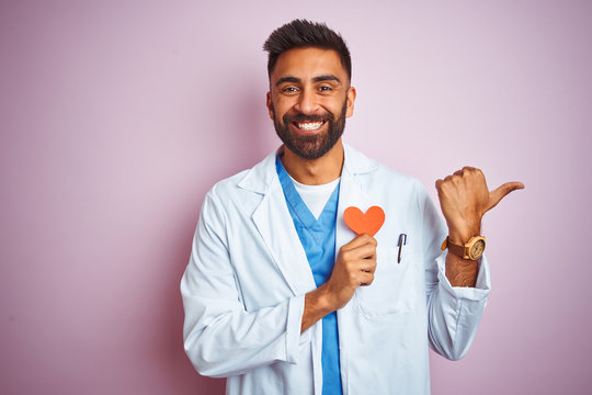 Young Indian Doctor Man Holding Paper Heart Standing Over Isolated Pink Background Pointing And Showing With Thumb Up To The Side With Happy Face Smiling