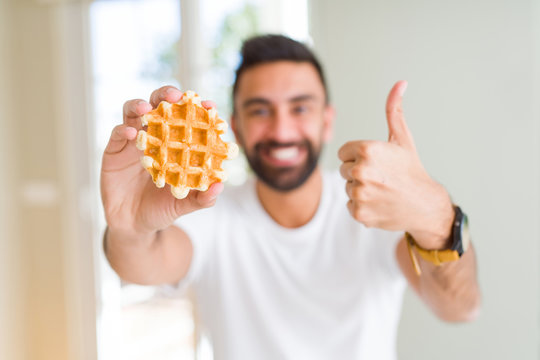 Handsome hispanic man eating sweet belgian waffle pastry happy with big smile doing ok sign, thumb up with fingers, excellent sign