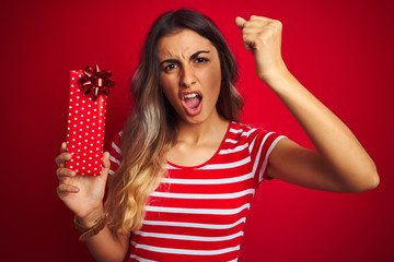Young beautiful woman holding birthday present over red isolated background annoyed and frustrated shouting with anger, crazy and yelling with raised hand, anger concept
