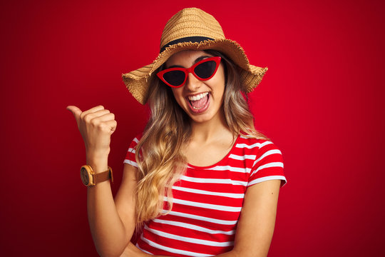 Young beautiful woman wearing sunglasses and summer hat over red isolated background smiling with happy face looking and pointing to the side with thumb up.