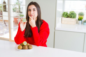 Beautiful young woman eating half fresh green kiwi serious face thinking about question, very...
