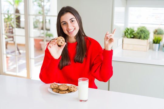 Beautiful young woman eating chocolate chips cookies and glass of milk very happy pointing with hand and finger to the side