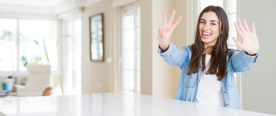 Wide angle picture of beautiful young woman sitting on white table at home showing and pointing up with fingers number eight while smiling confident and happy.