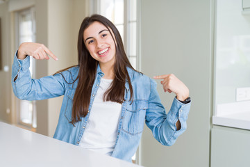 Beautiful young woman sitting on white table at home looking confident with smile on face, pointing...