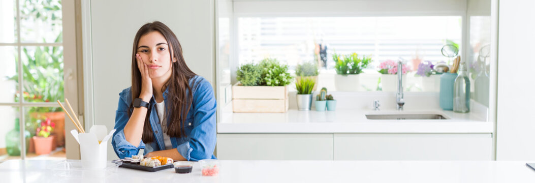 Wide angle picture of beautiful young woman eating delivery sushi thinking looking tired and bored with depression problems with crossed arms.