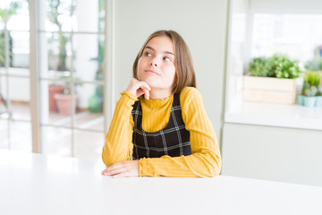 Beautiful young girl kid sitting on the table with hand on chin thinking about question, pensive expression. Smiling with thoughtful face. Doubt concept.