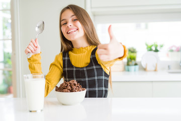 Beautiful young girl kid eating chocolate cereals and glass of milk for breakfast happy with big...