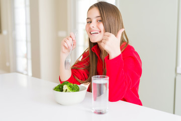 Beautiful young girl kid eating fresh broccoli and drinking water happy with big smile doing ok sign, thumb up with fingers, excellent sign