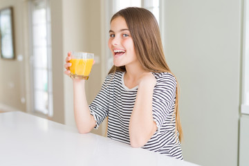 Beautiful young girl kid drinking a glass of fresh orange juice with surprise face pointing finger to himself