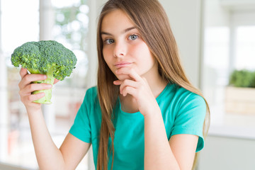 Beautiful young girl eating fresh broccoli serious face thinking about question, very confused idea