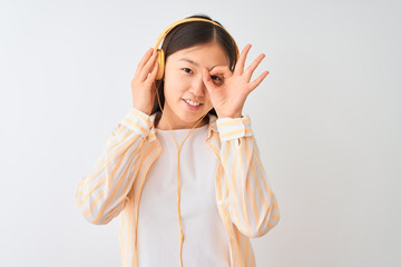 Young chinese woman listening to music using headphones over isolated white background with happy face smiling doing ok sign with hand on eye looking through fingers