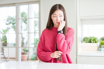 Beautiful Asian woman wearing pink sweater on white table looking stressed and nervous with hands on mouth biting nails. Anxiety problem.